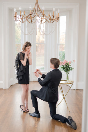 groom on one knee proposing to surprised bride to be wearing designer little black dress with one shoulder