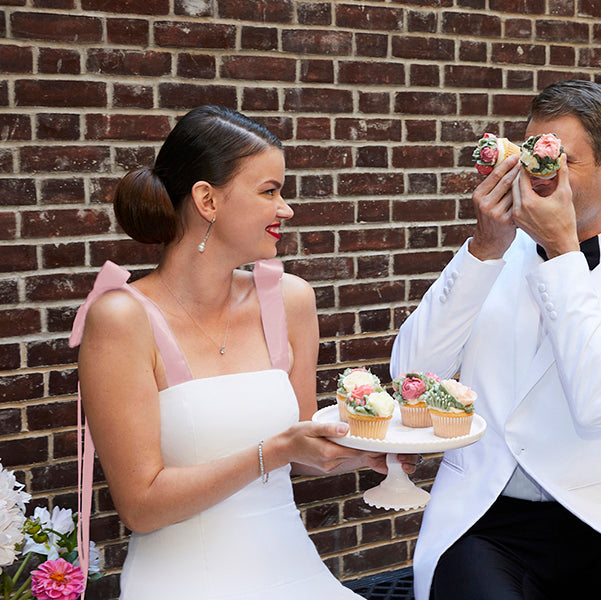 Beautiful bride wearing short white wedding dress with blush pink ribbon straps with groom and cupcakes