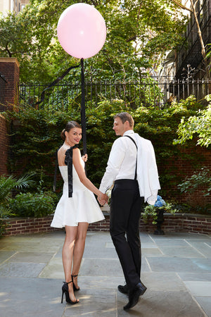 Bride walking in chic white cocktail dress with ribbons holding a pink balloon with her groom